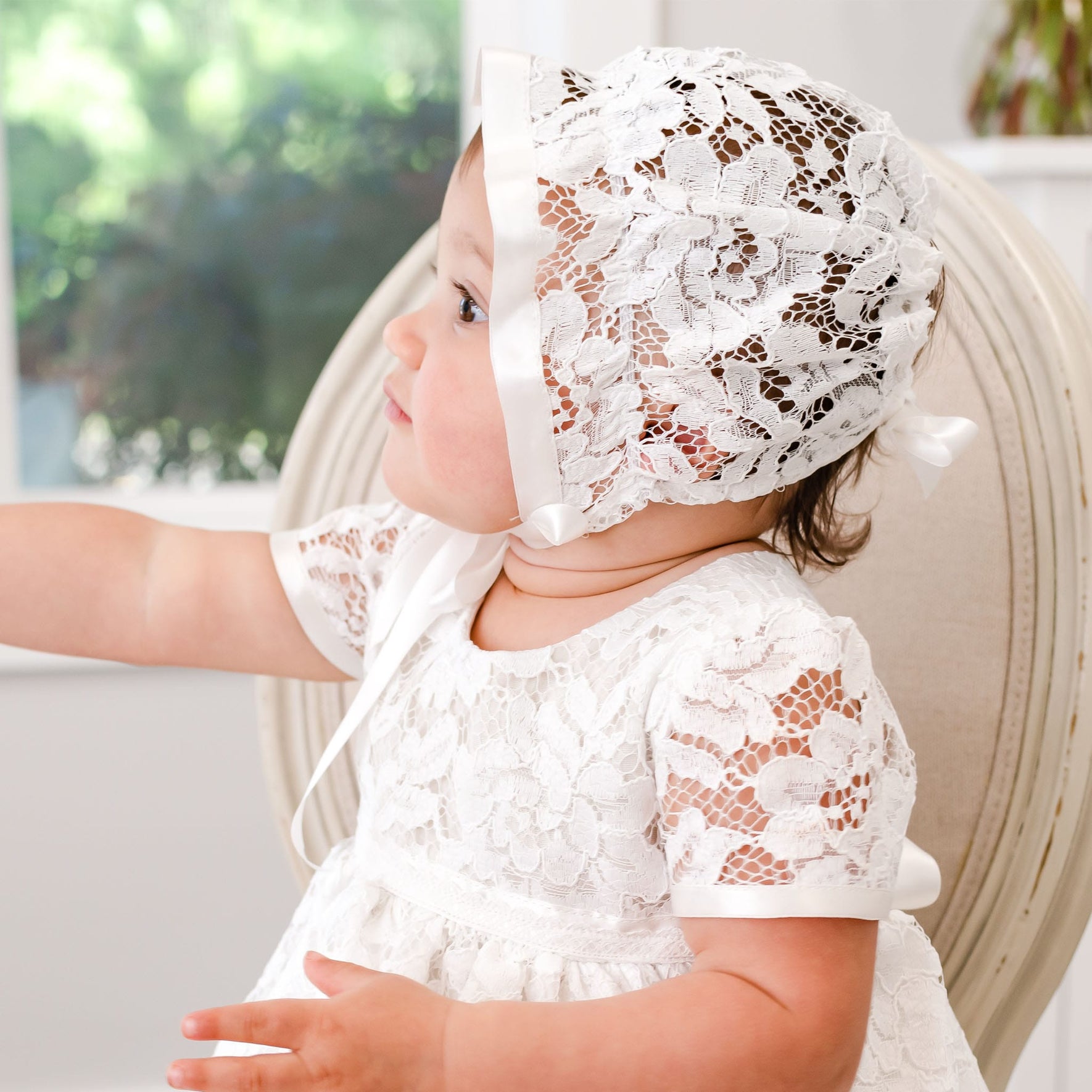 A baby with light skin wearing a Rose Christening Gown & Bonnet sits in a chair, gazing to the side in a brightly lit room.