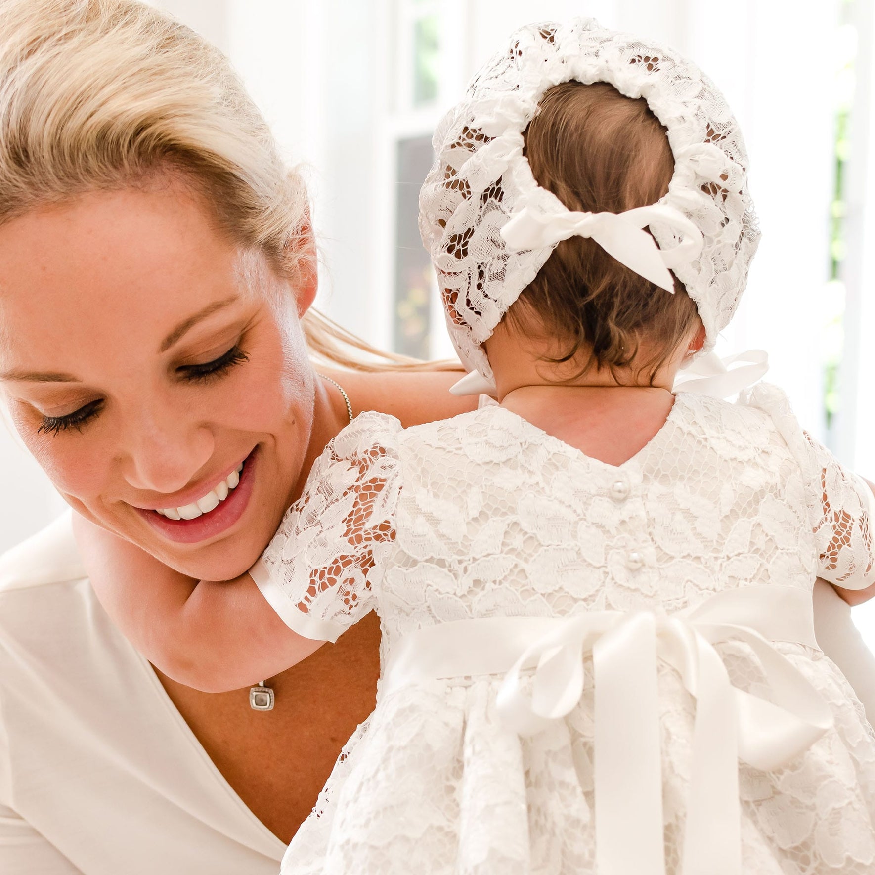 A mother, smiling, holds her baby dressed in a Rose Christening Gown & Bonnet. The baby's back is turned towards the camera, and they are in a brightly lit room.