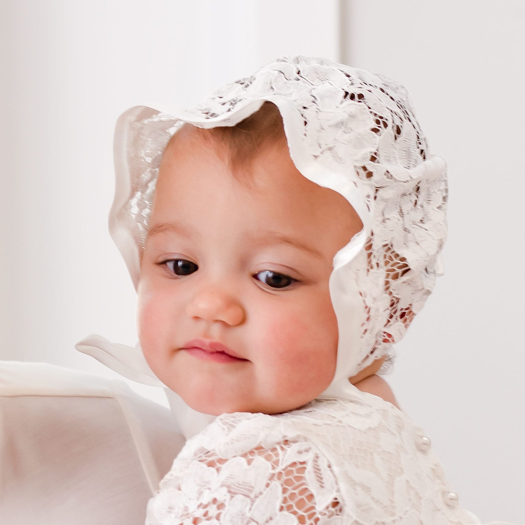 Close-up of a baby girl in the Rose Lace Christening Bonnet, crafted from fine ivory lace and accented with satin edging. She gazes off to the side with a calm expression, showcasing the bonnet’s beautiful lacework paired with her matching christening gown.
