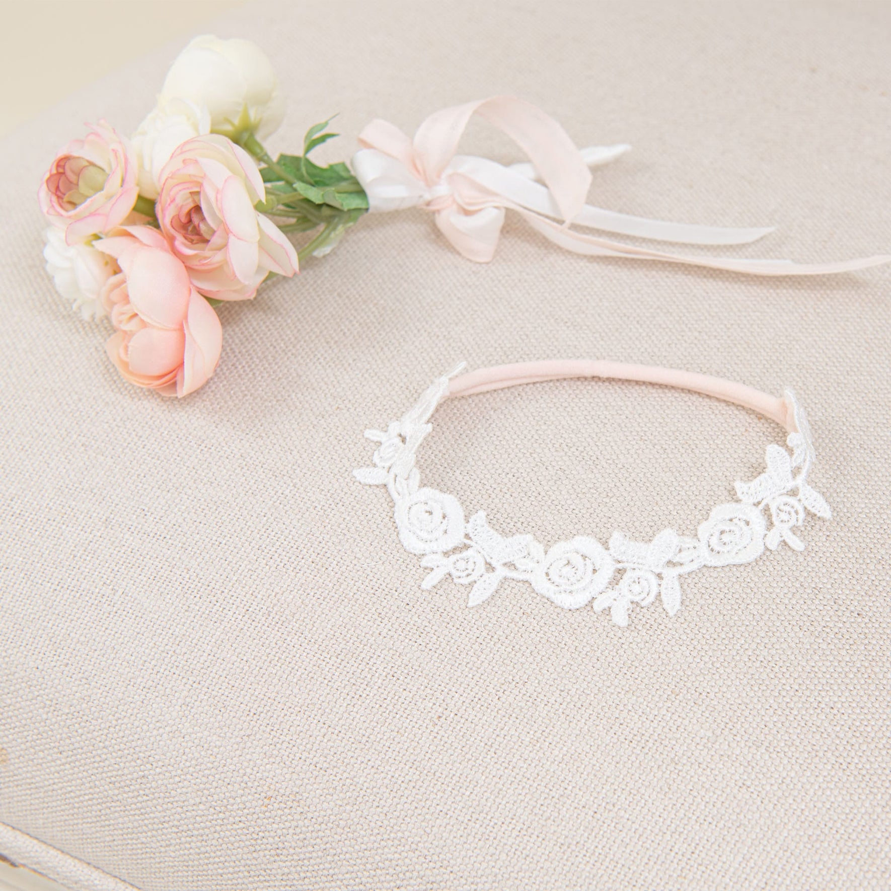 A Rose Lace Headband, adorned with delicate handcrafted lace roses, is displayed on a beige textured surface. Beside it rests a small bouquet of pink and white flowers tied with a ribbon, all softly illuminated to highlight their intricate features.