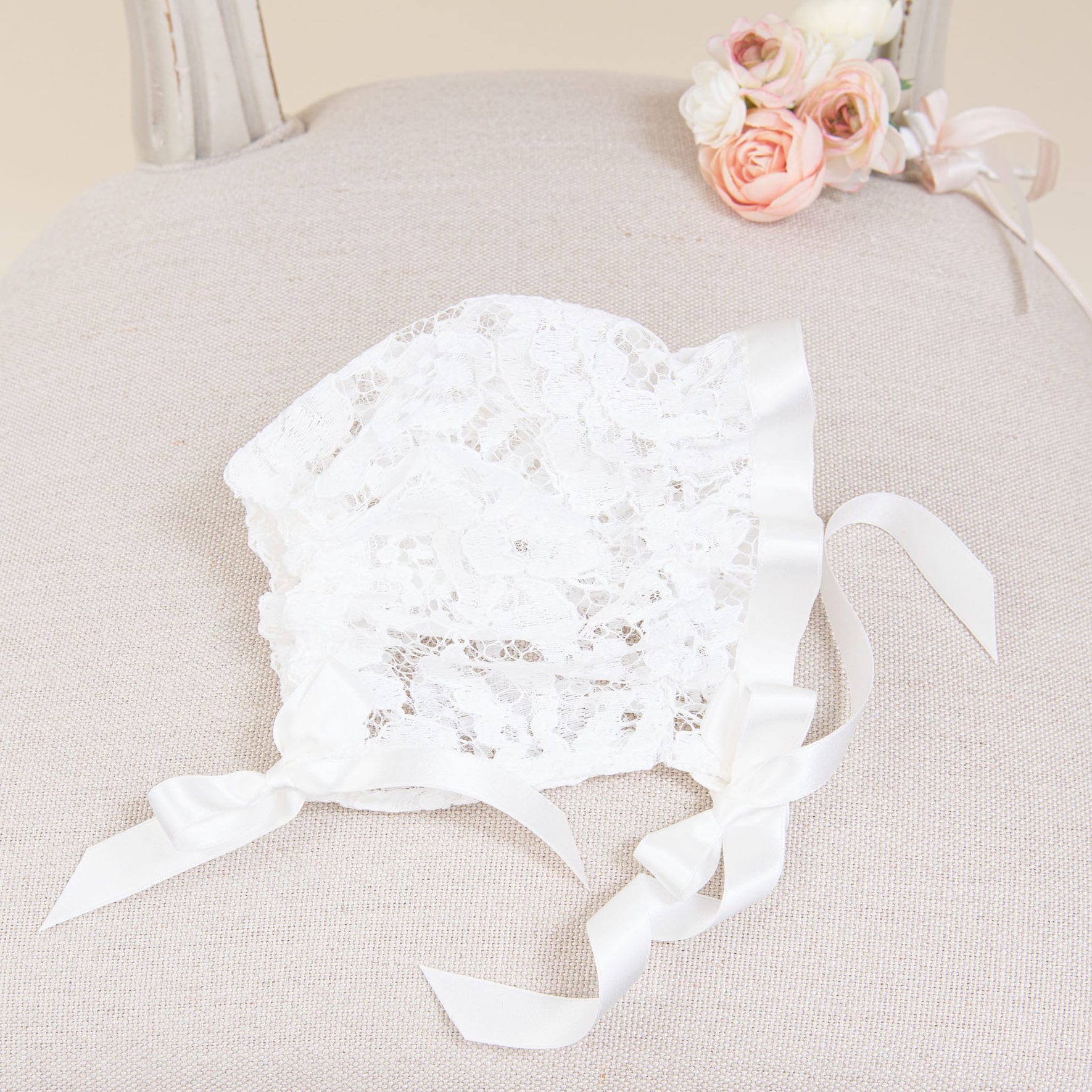A Rose Bonnet adorned with satin ribbons and bows rests on a beige cushioned chair. Next to it, there is a small floral bouquet featuring pink and white roses.