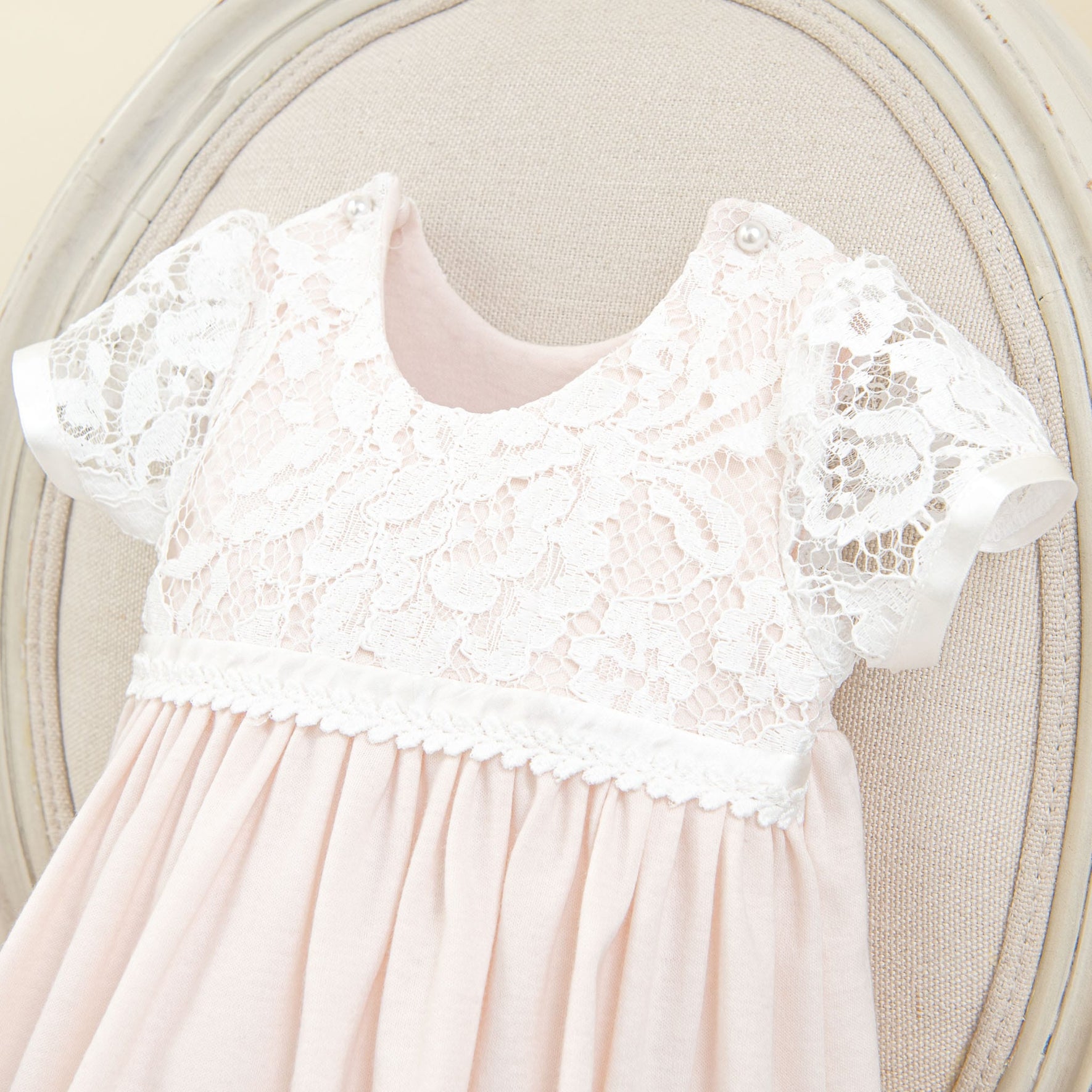 A pink baby dress with a white lace overlay on the top part of the dress and sleeves. The Rose Layette & Bonnet, part of a charming newborn layette, has scalloped edges on the lace and a band of white trim around the waist. It's displayed on a beige cushioned chair, perfect to pair with a Rose lace bonnet.