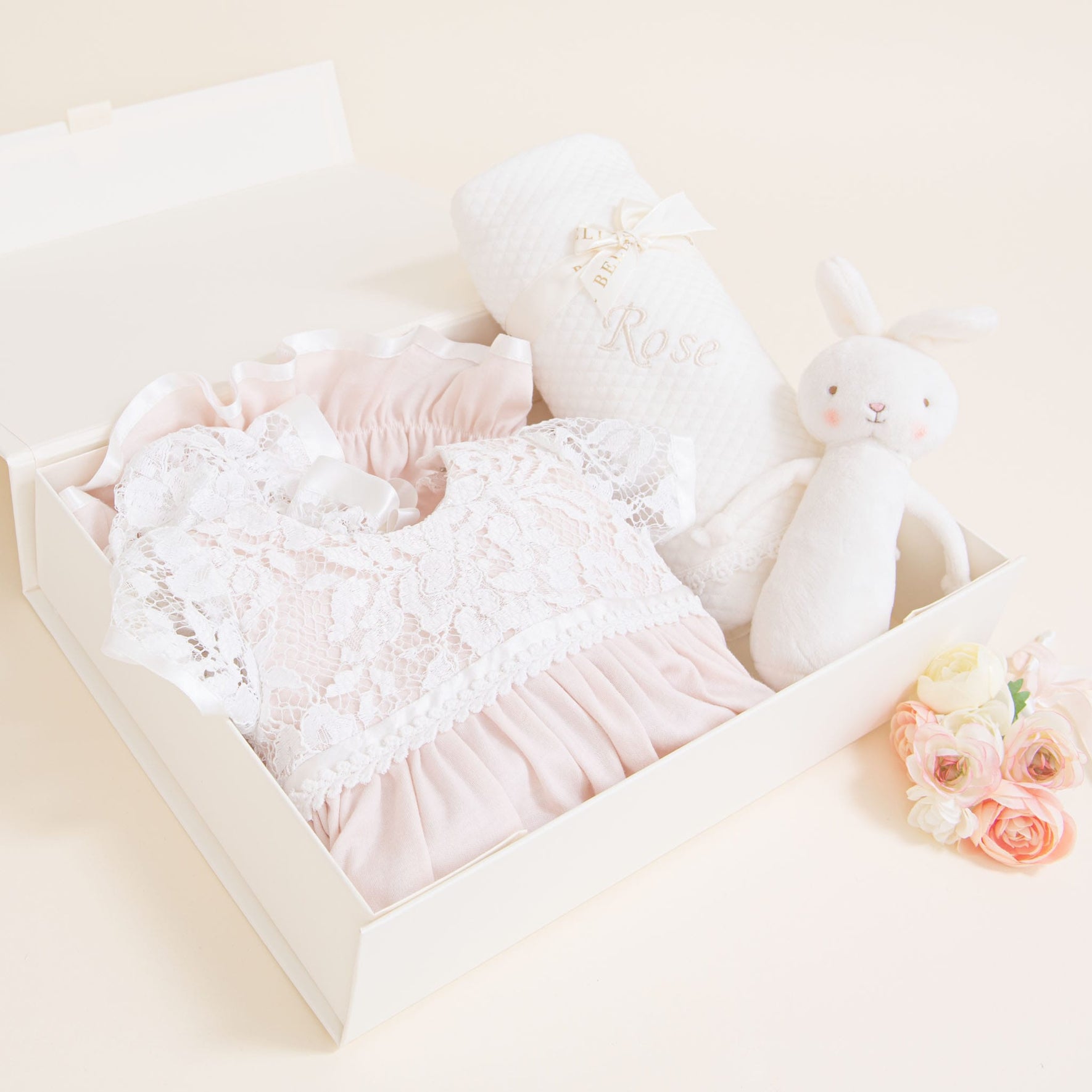 The Rose Newborn Gift Set - Save 10% contains a pink lace baby dress, a white baby blanket made of pima cotton with the name "Rose" embroidered, and a white stuffed bunny toy. Beside the box, there is a small bouquet of pink and white flowers.