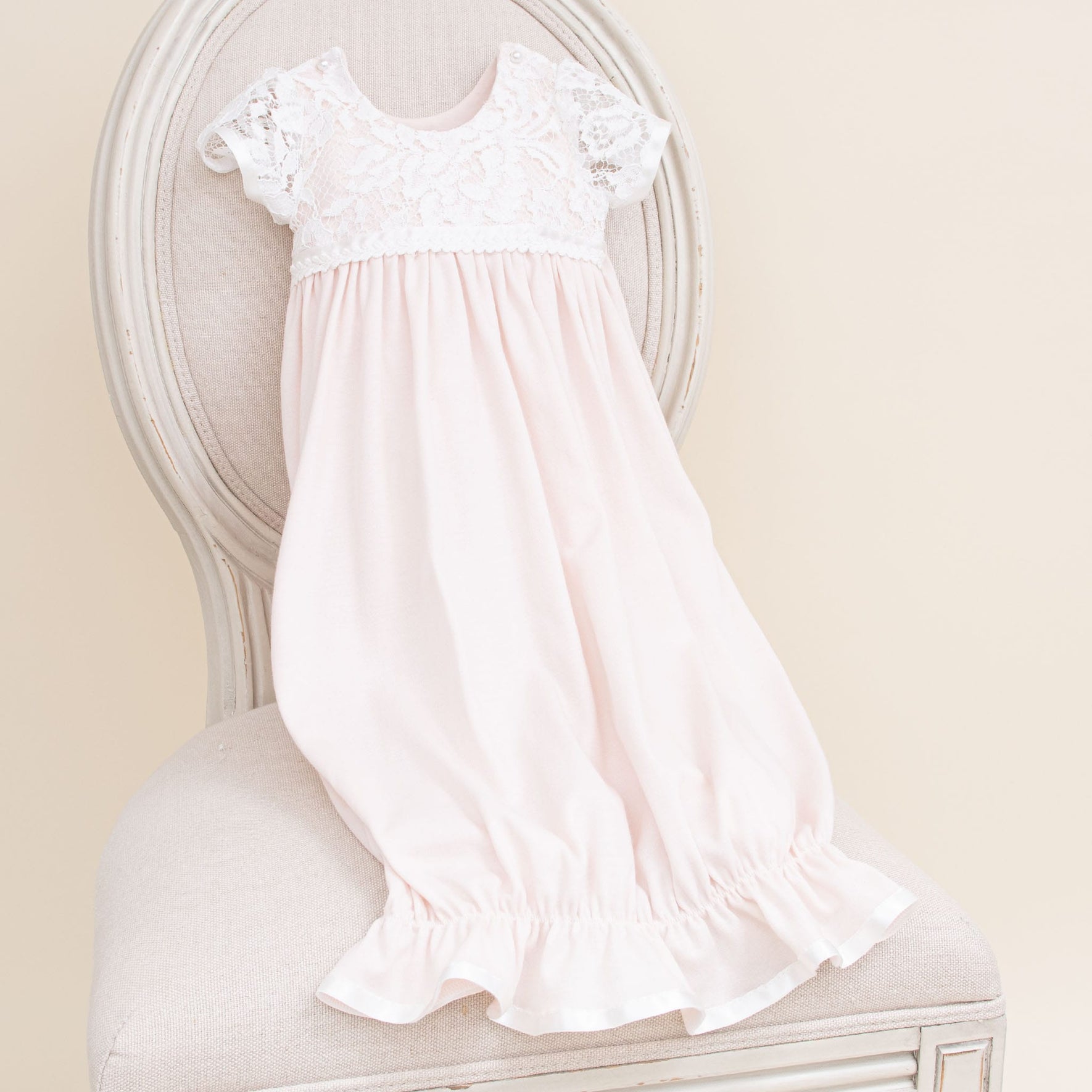 A soft pink Rose Newborn Gift Set - Save 10% with short rose-patterned lace sleeves and a ruffled hem is draped over the back of a light-colored, upholstered chair with a round backrest. The set features intricate lace detailing on the upper part and gentle, flowing pima cotton fabric below.