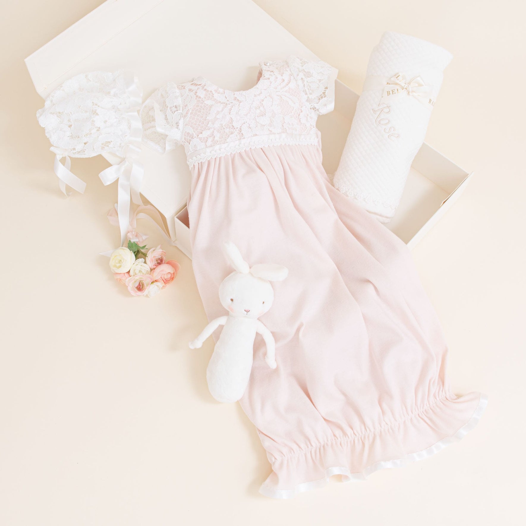 Rose Newborn Gift Set - Save 10% displayed in a box. Includes a pale pink newborn coming home gown with a rose patterned lace bodice, matching bonnet, white plush bunny toy, quilted blanket, and a small bunch of pastel-colored roses. The box is open on a light beige surface.