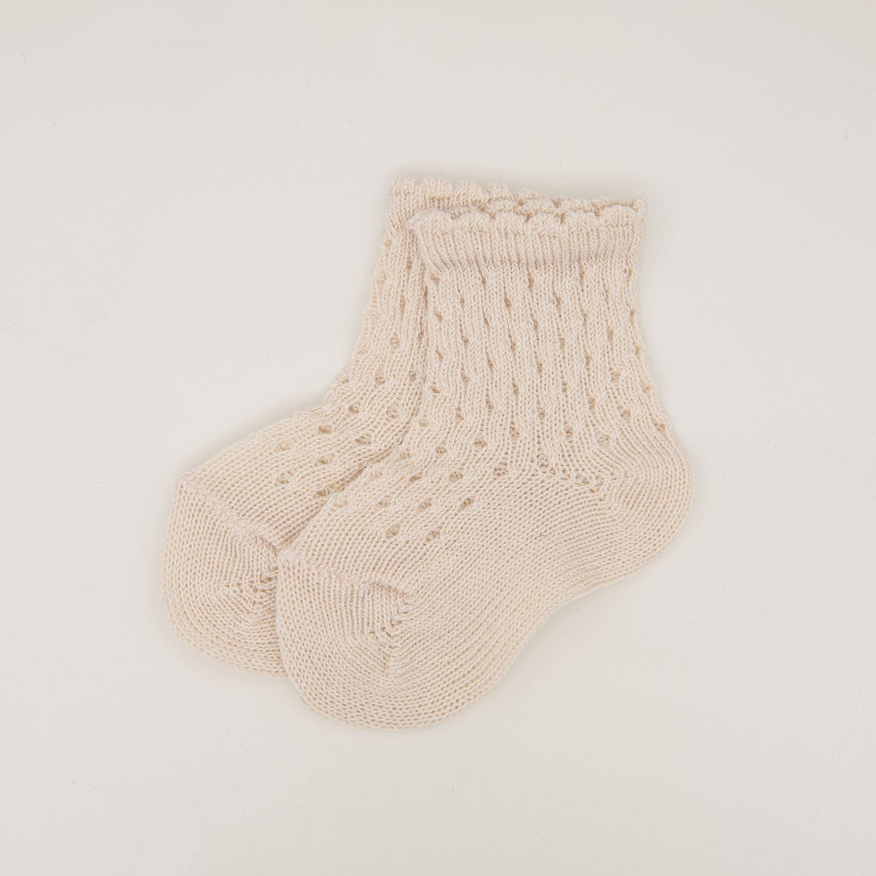 The Scallop Edge Crochet Sock, a pair of cream-colored knitted baby socks featuring a textured pattern and scalloped cuffs, is displayed against a plain white background. Made from 100% cotton, these delicate socks provide both comfort and style for little feet.