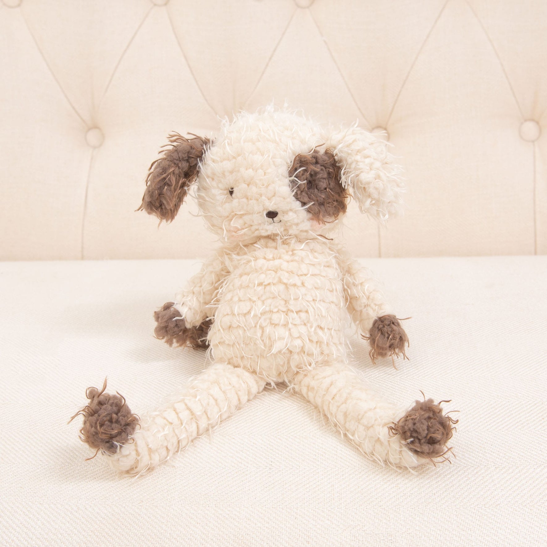 The Shaggy Fetch Puppy, a crocheted stuffed animal dog with a light cream body and dark brown ears, paws, and tail, sits on a beige tufted background. This plush shaggy puppy has an embroidered face, making it both baby-safe and adorable for little ones.