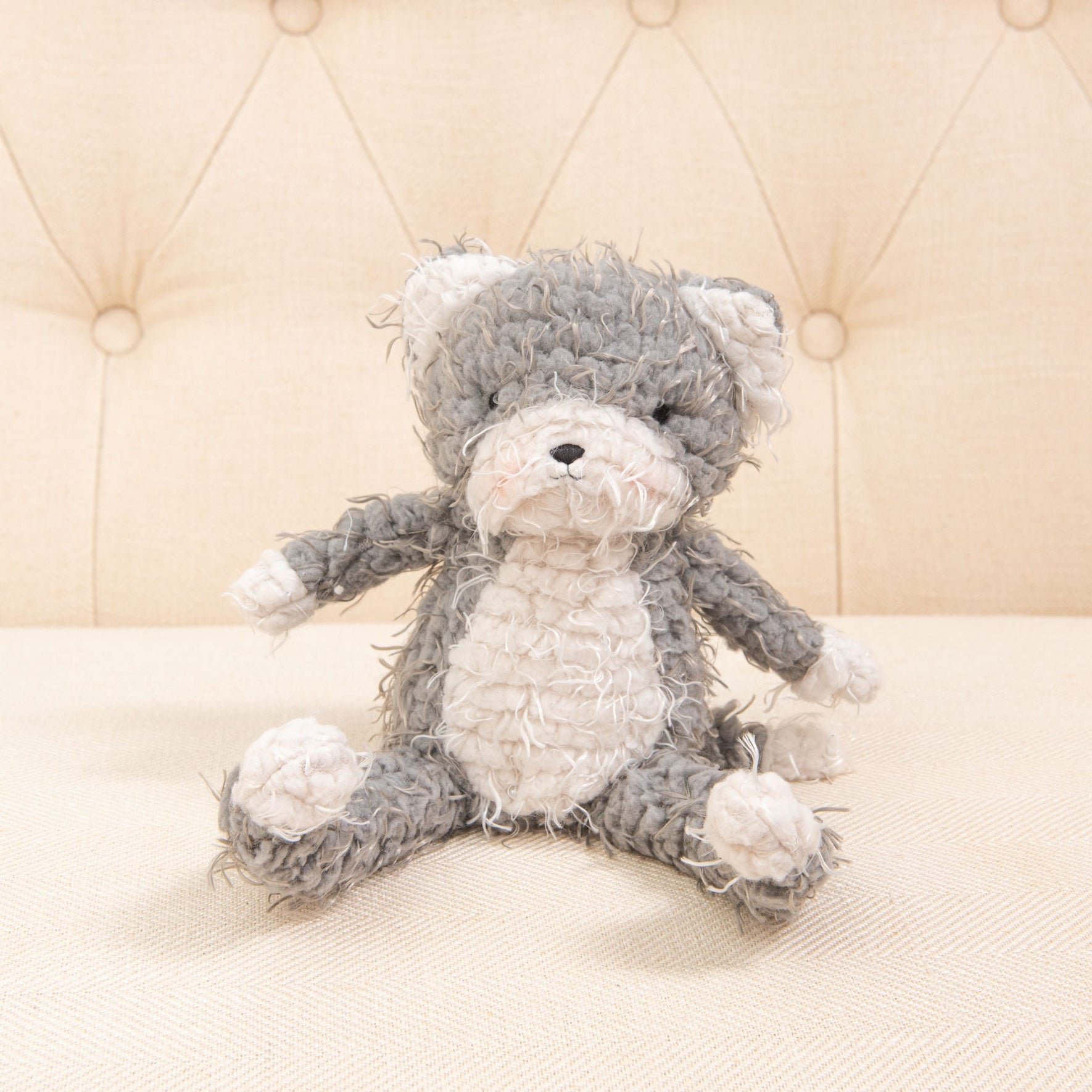 The Shaggy Purrty Kitty, in gray and white, rests on a beige tufted cushion with its embroidered face ensuring it's safe for babies.