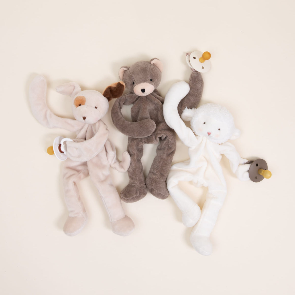 Arranged on a light surface are three Silly Buddy plush animal toys. From left to right, you'll find a beige dog, a brown bear, and a white sheep. Each of these cuddly companions features a pacifier clip in its left hand and is crafted from soft velour for heartwarming comfort.