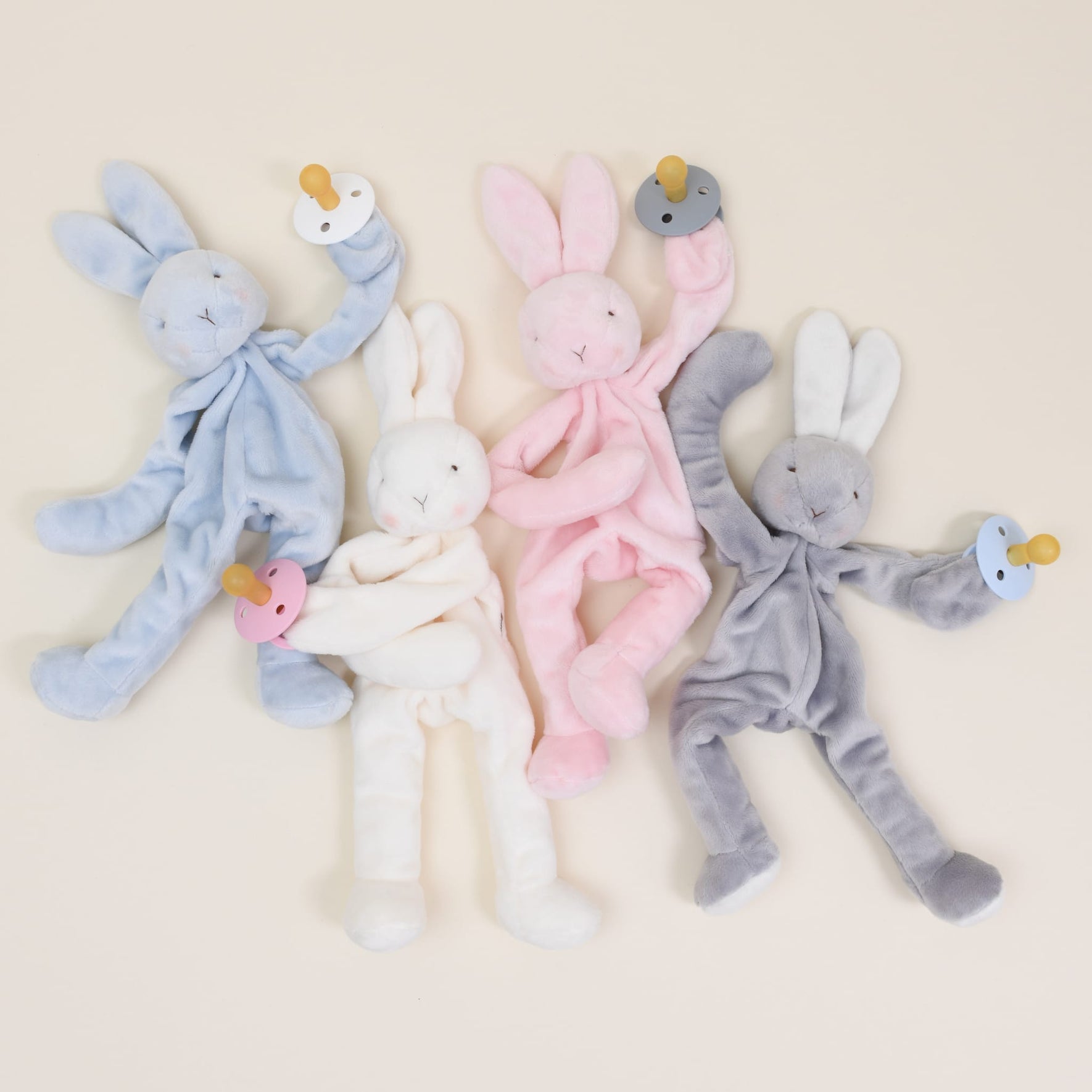 Four Silly Bunny Buddy Pacifier Holders in blue, white, pink, and gray are arranged on a neutral background. Each baby-safe rabbit plush features floppy ears and limbs while holding a matching colored pacifier with a clip: blue, white, pink, and gray.