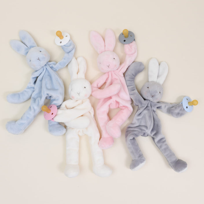 Four Silly Bunny Buddy Pacifier Holders in blue, white, pink, and gray are arranged on a neutral background. Each baby-safe rabbit plush features floppy ears and limbs while holding a matching colored pacifier with a clip: blue, white, pink, and gray.