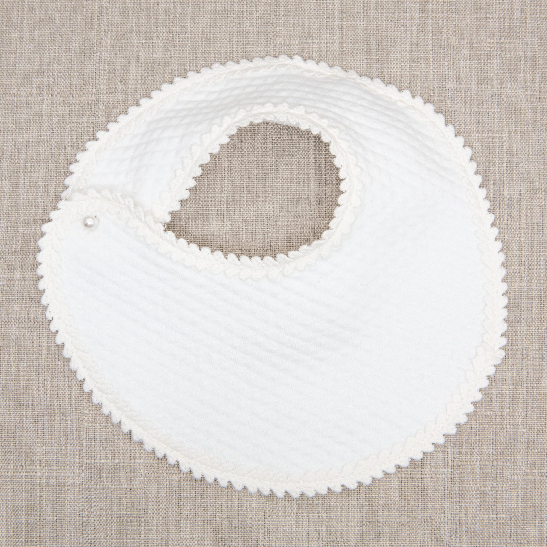 Flat lay photo of the Tessa Quilt Bib. The bib is made with a soft quilted cotton, accented with a floral edge lace, and features a pearl style button closure.