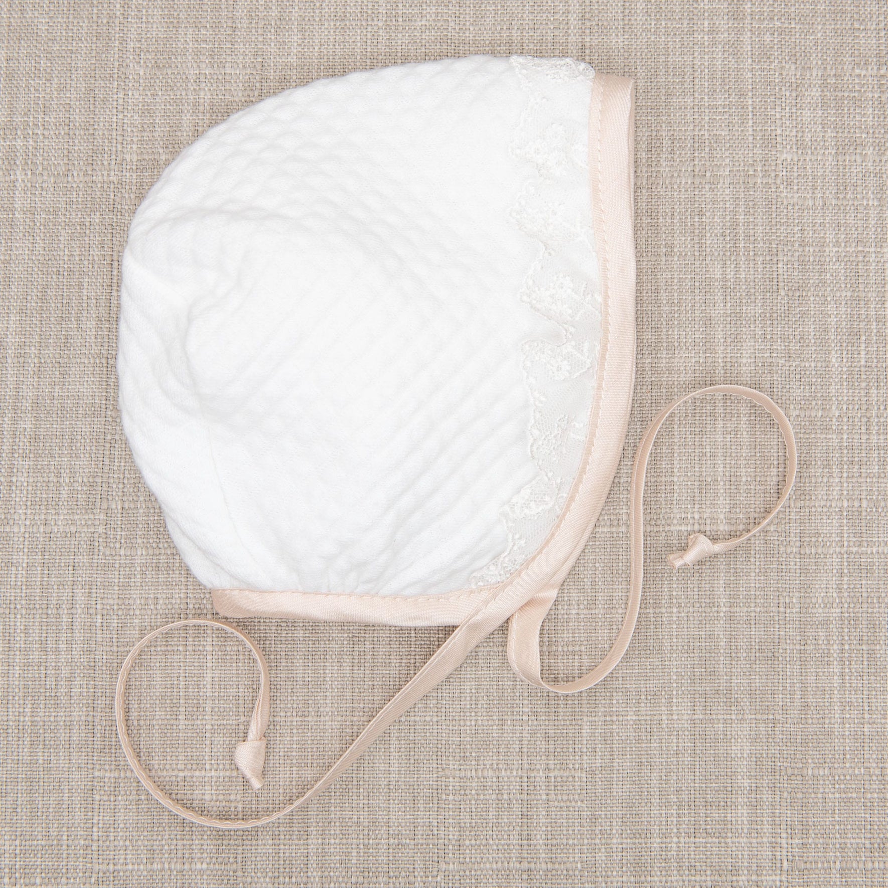 Flat lay photo of the Tessa Quilt Bonnet displayed on a beige fabric surface. The angle showcases the soft quilted cotton and trim in pink silk and ivory floral lace.