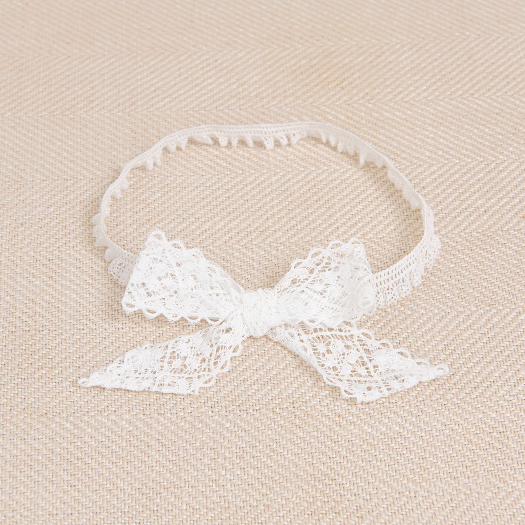 The Thea Lace Headband, handcrafted in the USA, is beautifully displayed on a beige textured surface. Its stretch lace features a delicate, intricate pattern, and the bow is neatly tied at the front.