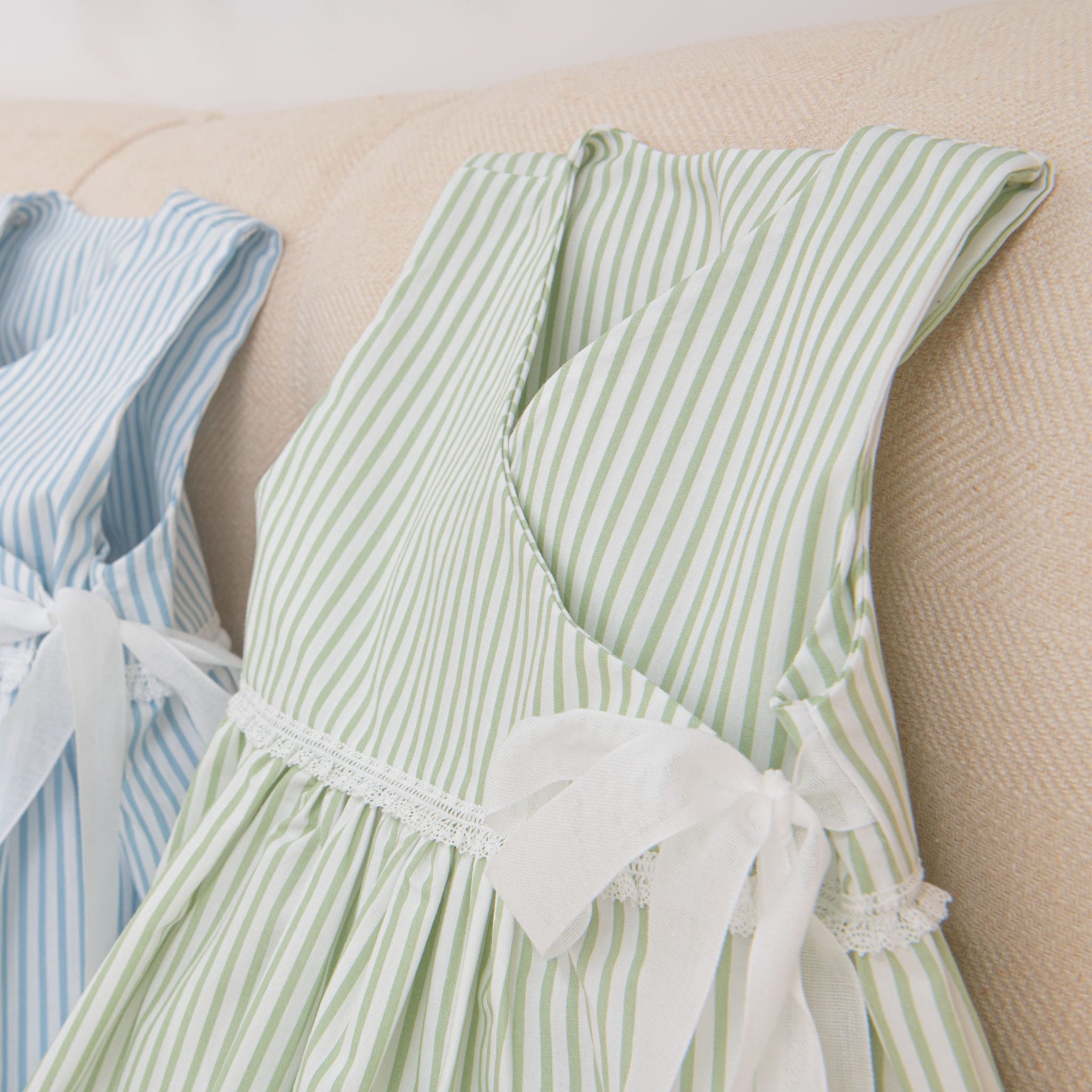 On a beige surface lie two striped cotton dresses placed side by side. The first is Thea Wrap Dress, showcasing green and white vertical stripes adorned with a white ribbon, while the second dress features blue and white stripes. Both dresses pair perfectly with an elegant Thea Bow Headband.