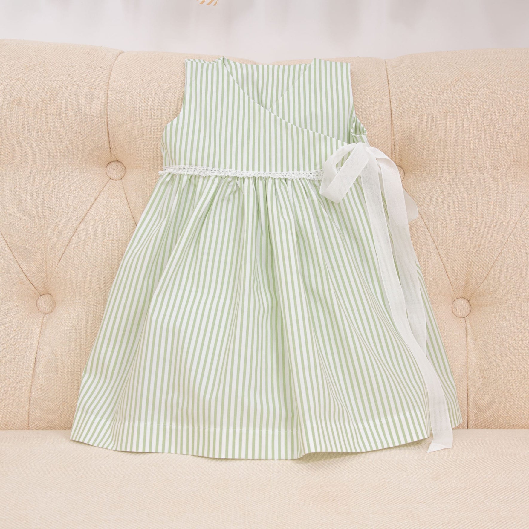 The Thea Wrap Dress, a sleeveless children's dress crafted from green and white striped cotton, features a charming white ribbon tied at the waist and is elegantly showcased on a light beige tufted cushion. It pairs beautifully with Thea Bow Headband.