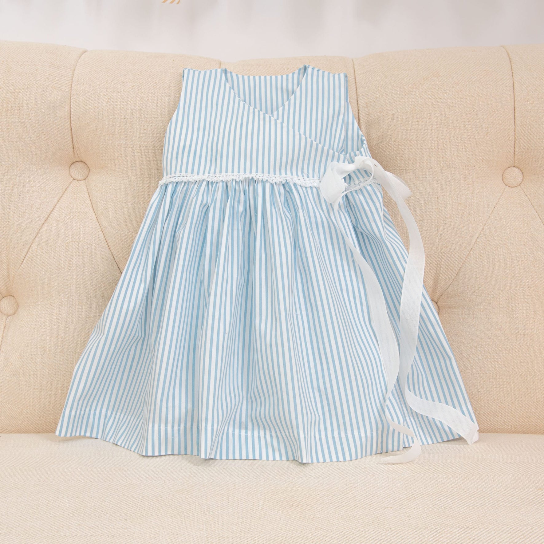 The Thea Wrap Dress, a children's cotton sleeveless outfit with light blue and white stripes, is displayed on a beige upholstered surface. It showcases a v-neckline and is adorned with a white ribbon bow at the waist, beautifully complementing the matching Thea Bow Headband.