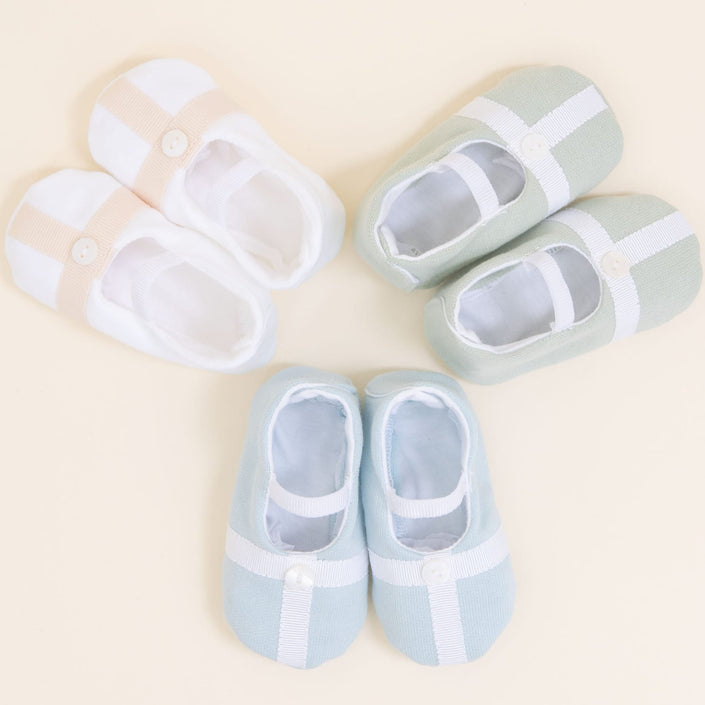 Flat lay photo showing the top detail of three pairs of Theodore Booties, including blue, green, and a white/tan color. The booties are made from a 100% French Terry Cotton with a Grosgrain Trim (with button detail) and soft elastic strap.