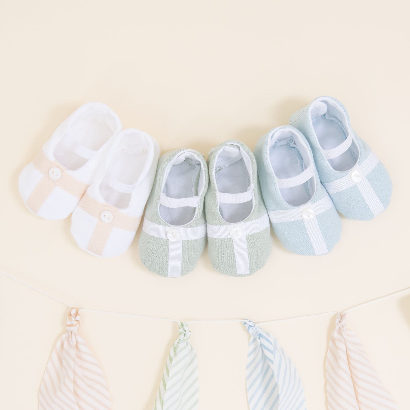 Flat lay photo showing the top detail of three pairs of Theodore Booties, including blue, green, and a white/tan color. The booties are made from a 100% French Terry Cotton with a Grosgrain Trim (with button detail) and soft elastic strap.