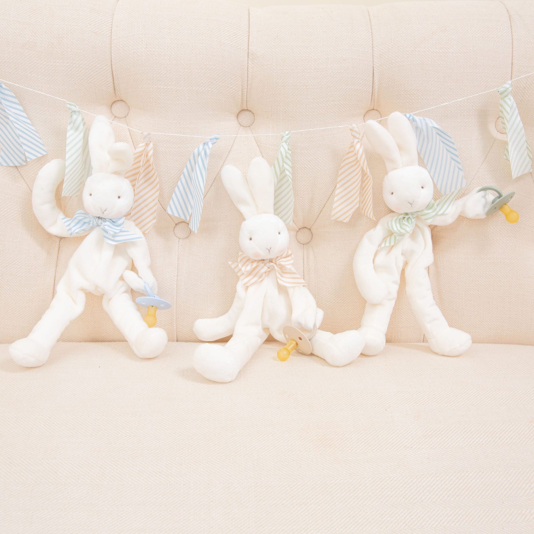 Flat lay photo of three Theodore Bunny Buddy Pacifier Holders. Stuffed animal floppy bunny made from a soft velour. Available colors include blue, green, and tan/white.