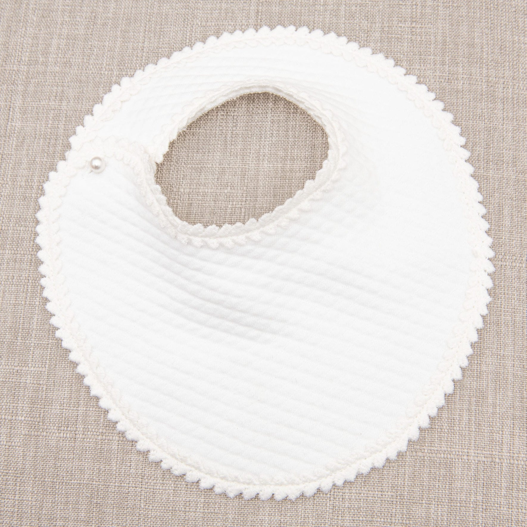 A Victoria Christening Bib is laid flat on a beige textured surface. The bib features a scalloped edge, a quilted texture, and a snap button closure at the neck.