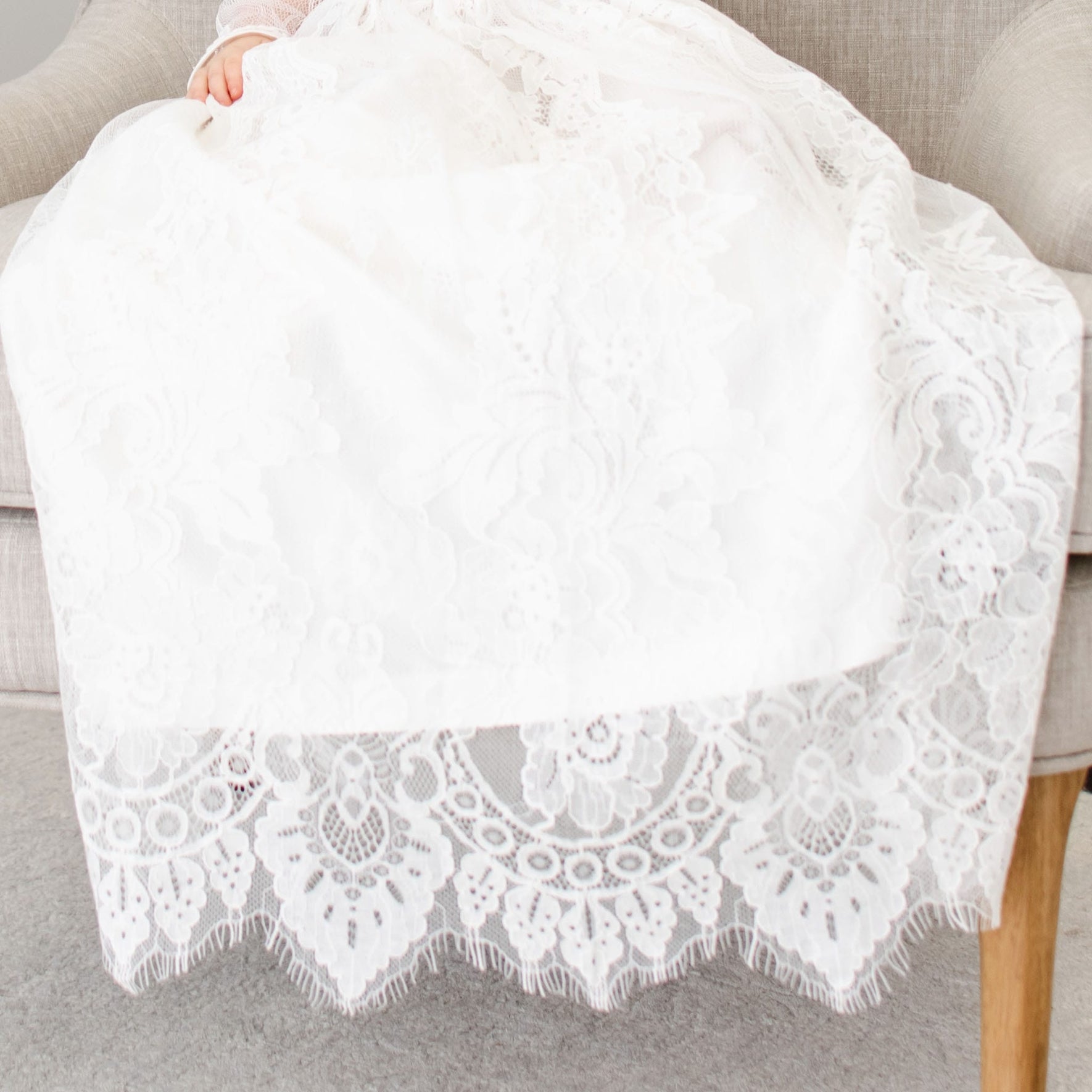 A child wearing the Victoria Long Sleeve Christening Gown & Bonnet is seated on a gray upholstered chair. The gown showcases intricate floral embroidery with a scalloped hem. The image focuses on the lower half of the child, emphasizing the ornate lace design.