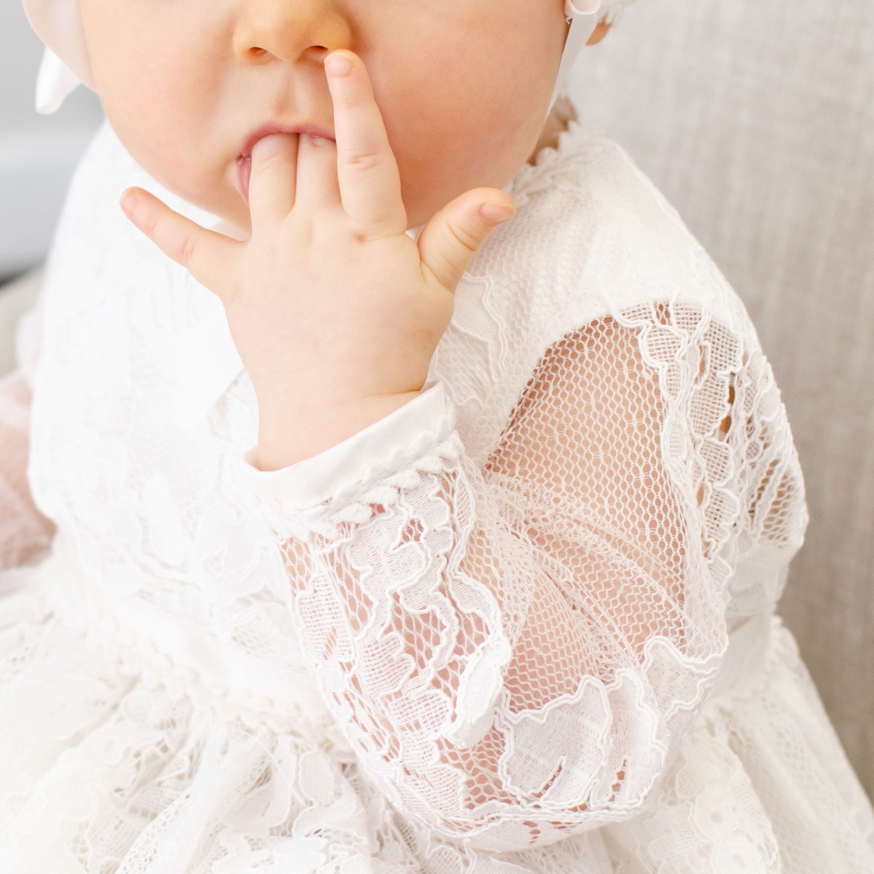 A baby in the Victoria Long Sleeve Christening Gown & Bonnet is sitting on a light-colored couch, fingers in their mouth. The white bonnet with a ribbon perfectly highlights the intricate lace details of the gown, capturing a timeless moment. Only part of the baby's face is visible, but the focus remains on this exquisite outfit.