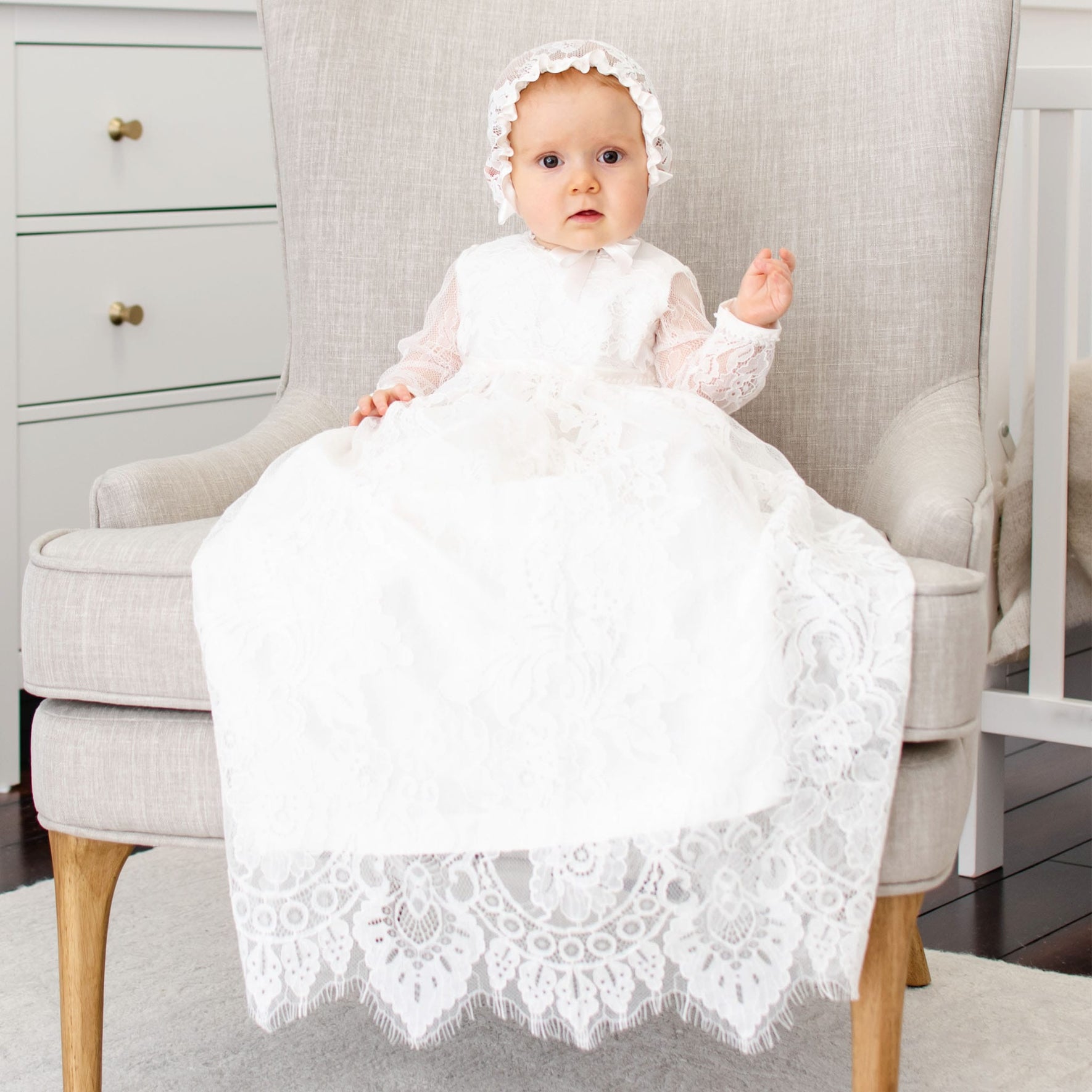 A baby is nestled on a light gray armchair, wearing the Victoria Long Sleeve Christening Gown & Bonnet, an exquisite outfit adorned with embroidered lace. The room features a dresser with two white drawers in the background, and the dark wood floor is partially covered by a light rug.