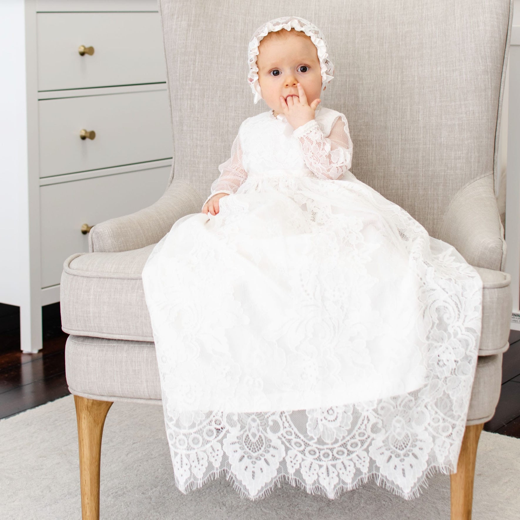 A baby dressed in the Victoria Long Sleeve Christening Gown & Bonnet sits on a light gray armchair. The baby has one hand near the mouth and looks towards the camera. The room is decorated with a white dresser featuring brass knobs and a white rug on a dark wood floor.