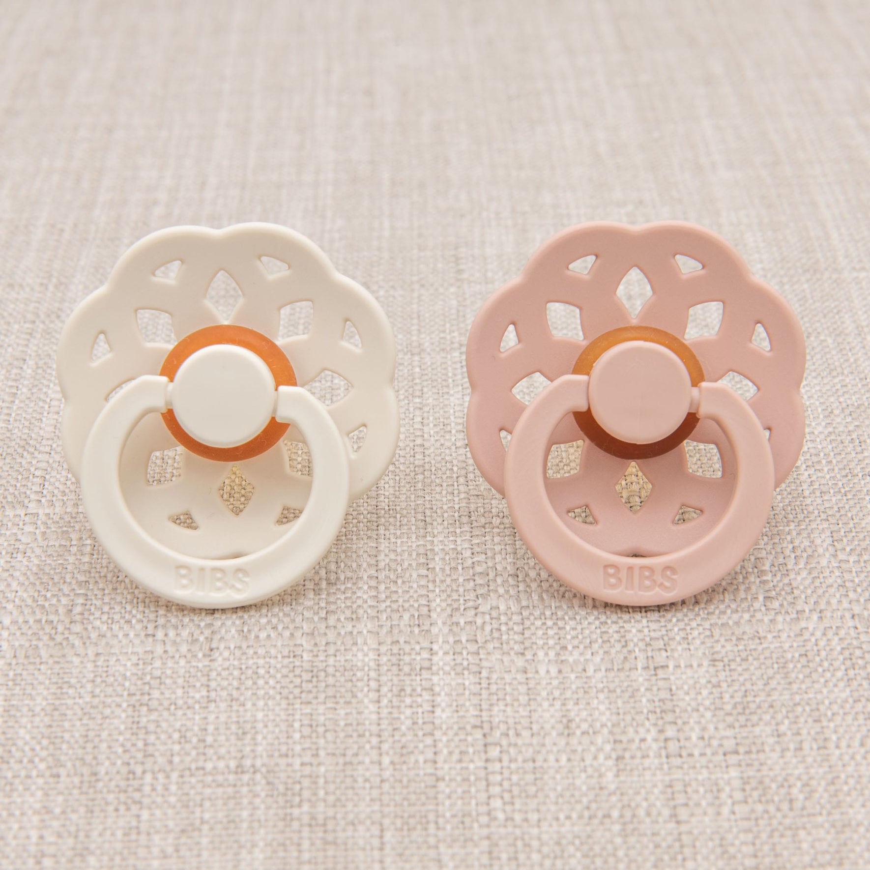A pair of Victoria pacifiers is displayed on a textured surface. Made in Denmark, one pacifier is ivory while the other is blush, both crafted from natural rubber to aid babies in self-soothing. Each features a circular shield and ring handle, with "BIBS" branding.