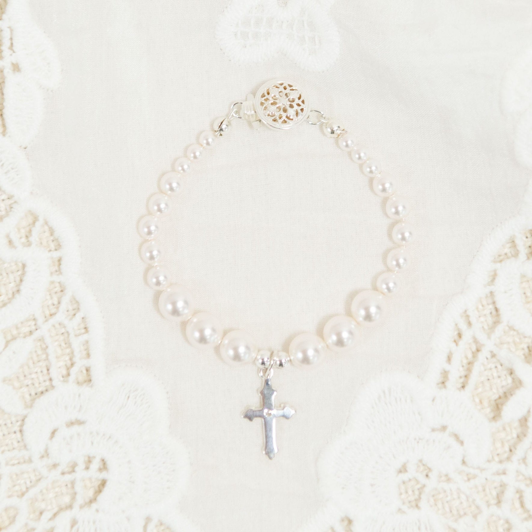 The White Luster Pearl Bracelet with Silver Cross is elegantly displayed on a white lace fabric background, making it an ideal Christening gift. The bracelet boasts a decorative circular clasp, enhancing its intricate beauty.