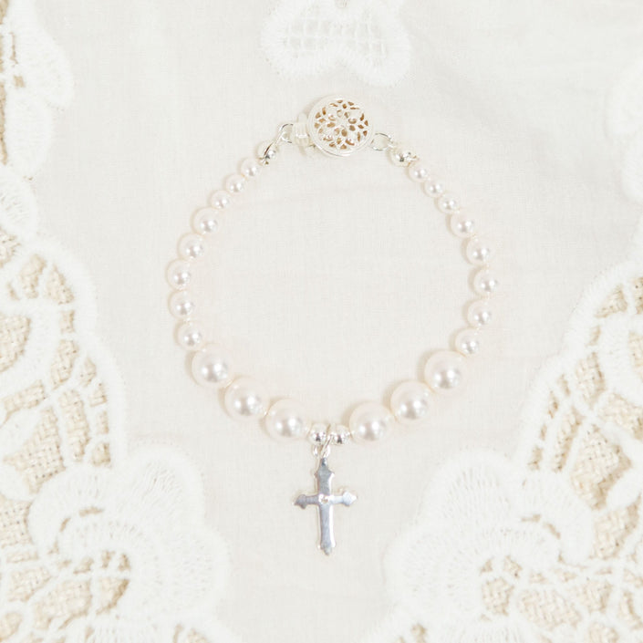 The White Luster Pearl Bracelet with Silver Cross is elegantly displayed on a white lace fabric background, making it an ideal Christening gift. The bracelet boasts a decorative circular clasp, enhancing its intricate beauty.