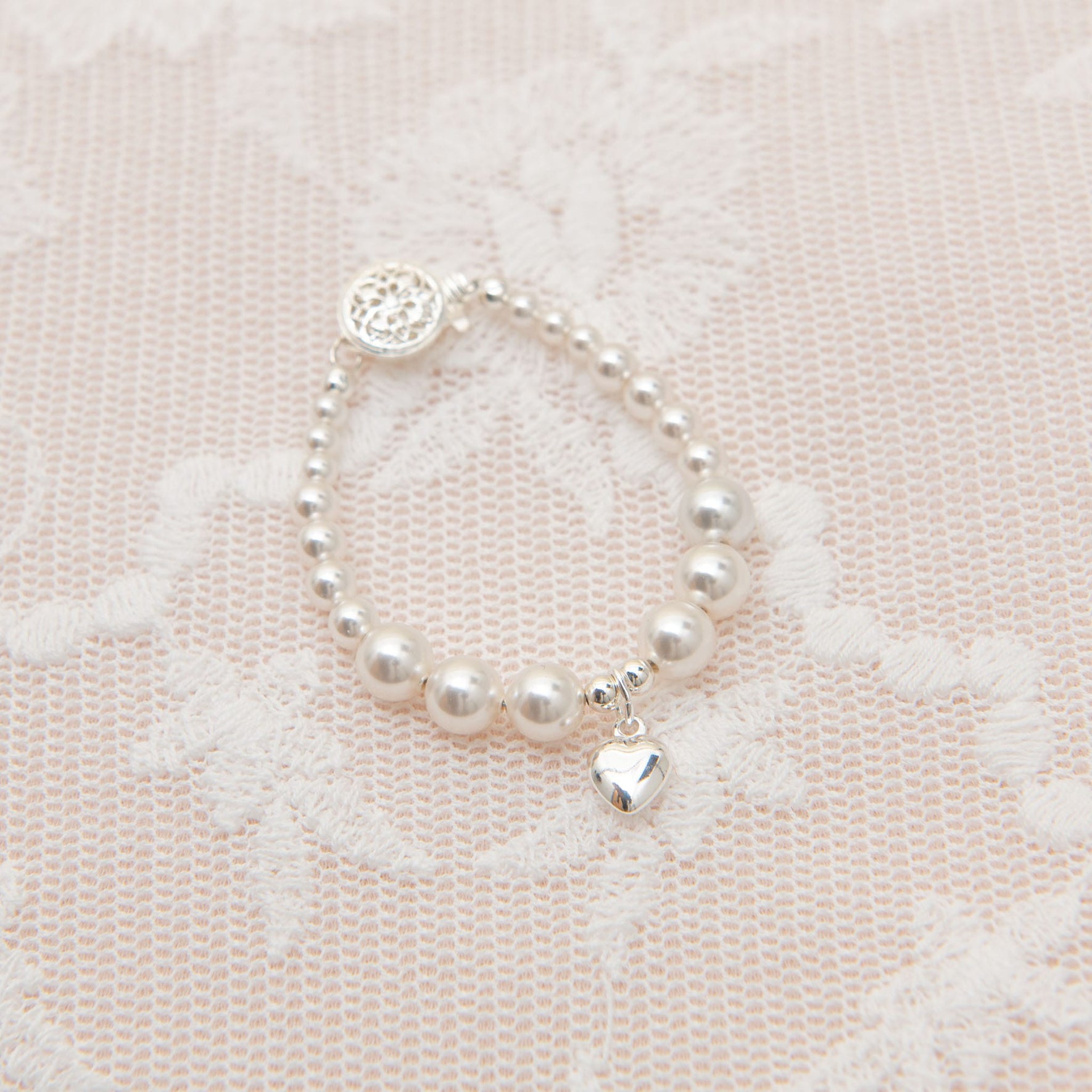 The White Luster Pearl Bracelet with Silver Heart Charm, featuring smooth round beads and a small sterling silver heart-shaped accent, is elegantly displayed on a delicate lace fabric background.