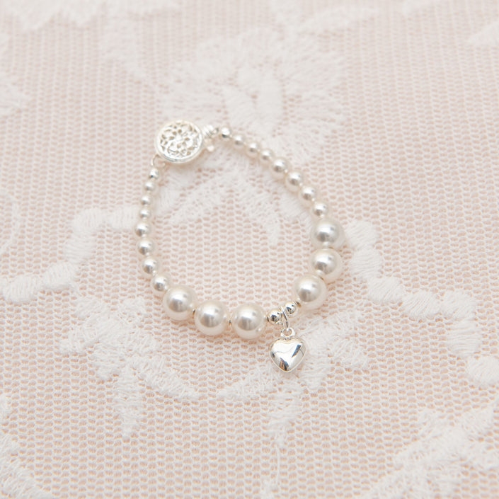 The White Luster Pearl Bracelet with Silver Heart Charm, featuring smooth round beads and a small sterling silver heart-shaped accent, is elegantly displayed on a delicate lace fabric background.