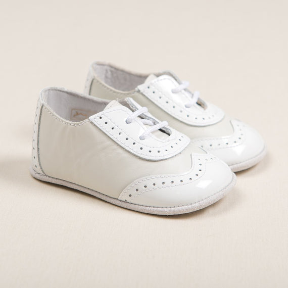 Flat lay photo of the Ivory Two Tone Wingtip Shoes that match the Oliver Baby Romper. They are made with a white patent leather and ivory matte leather with detailed edging. But bottoms are made with ivory suede leather.