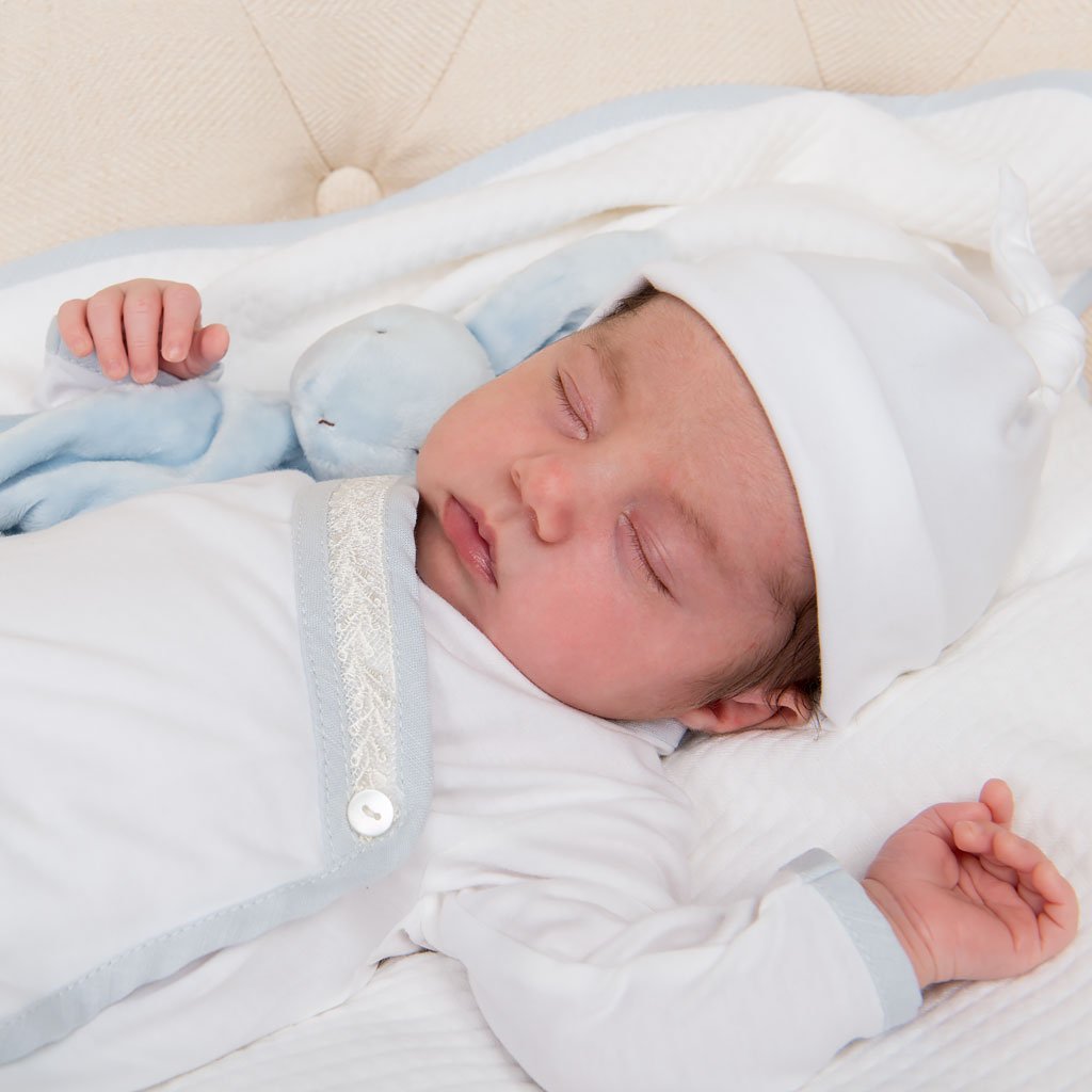 Close up detail of a newborn baby boy laying on a Harrison Personalized Blanket and holding a bunny stuffed animal. The boy is wearing the Harrison Knot Gown and Hat made with 100% pima cotton in white, detailed with a light blue cotton trim and an ivory Venice lace