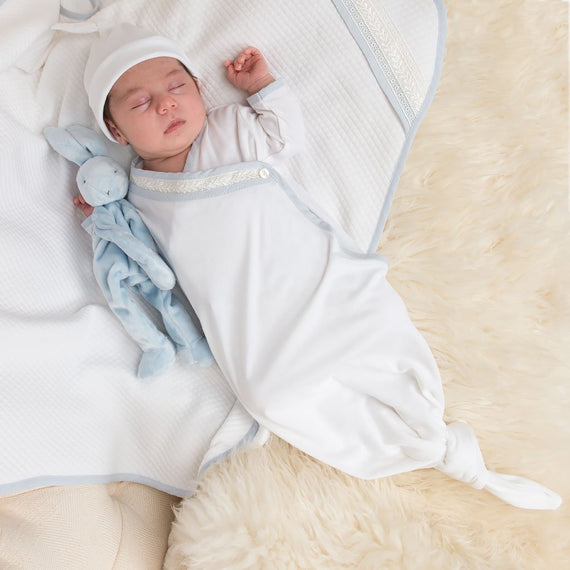 Newborn baby boy laying on a Harrison Personalized Blanket and holding a bunny stuffed animal. The boy is wearing the Harrison Knot Gown and Hat made with 100% pima cotton in white, detailed with a light blue cotton trim and an ivory Venice lace