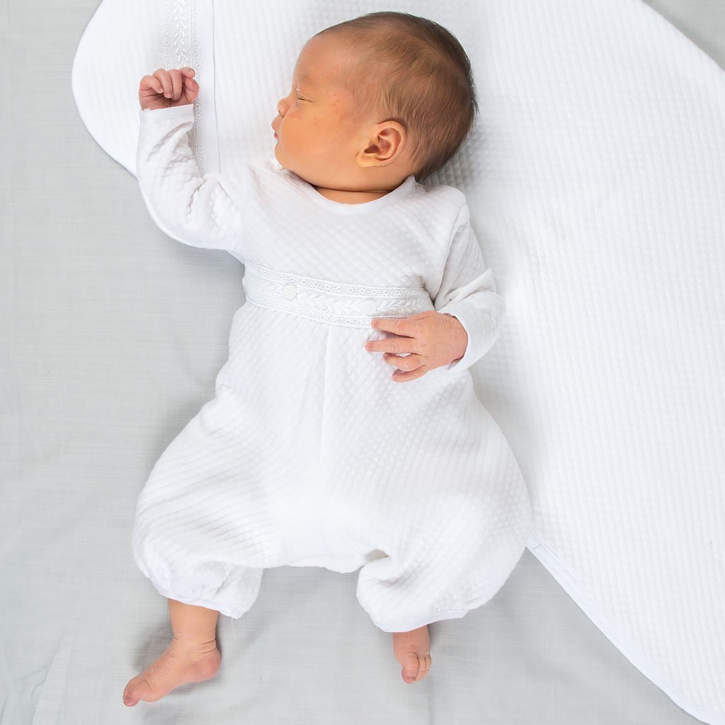 A newborn baby sleeps on a Elijah Personalized Blanket and wears the Elijah Newborn Romper made from a plush white quilted cotton, featuring white Venice lace at the bodice and white linen trim at the cuffs and neck