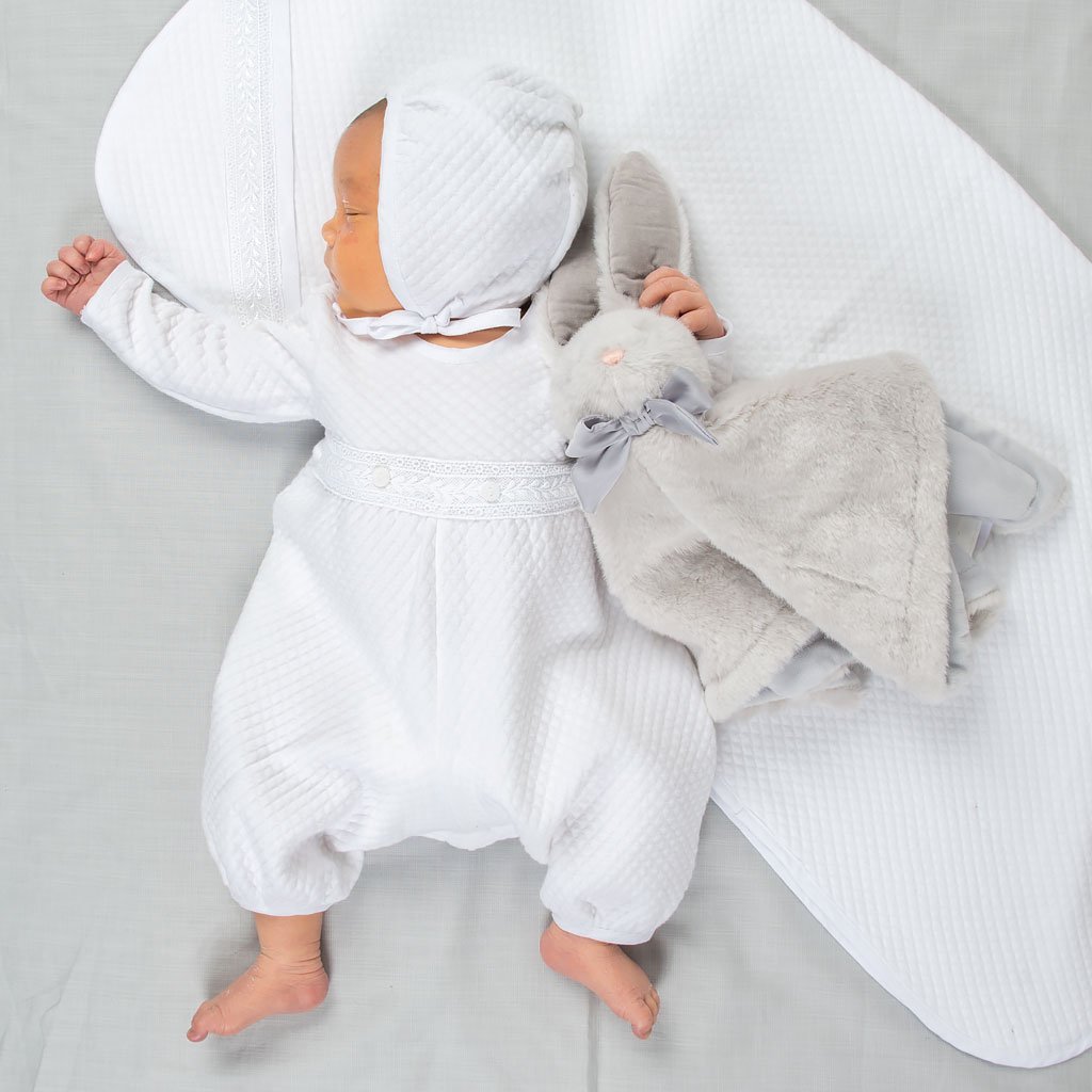 A newborn baby sleeps on a Elijah Personalized Blanket and wears the Elijah Newborn Romper made from a plush white quilted cotton, featuring white Venice lace at the bodice and white linen trim at the cuffs and neck. The baby is holding a stuffed bunny