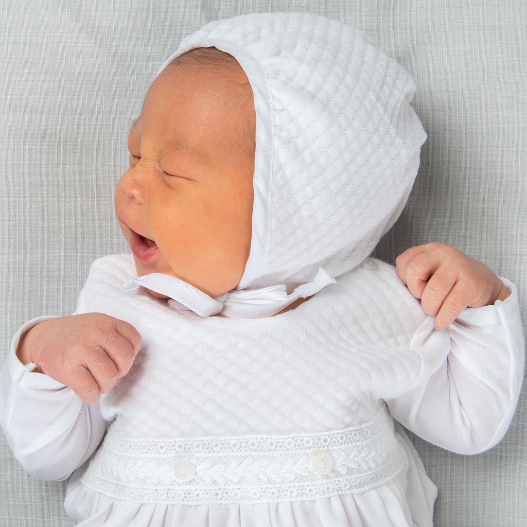 Newborn baby wearing an Elijah Newborn Quilted Bonnet made with soft quilted cotton in white and trimmed in white silk. White linen ties under the chin of the baby