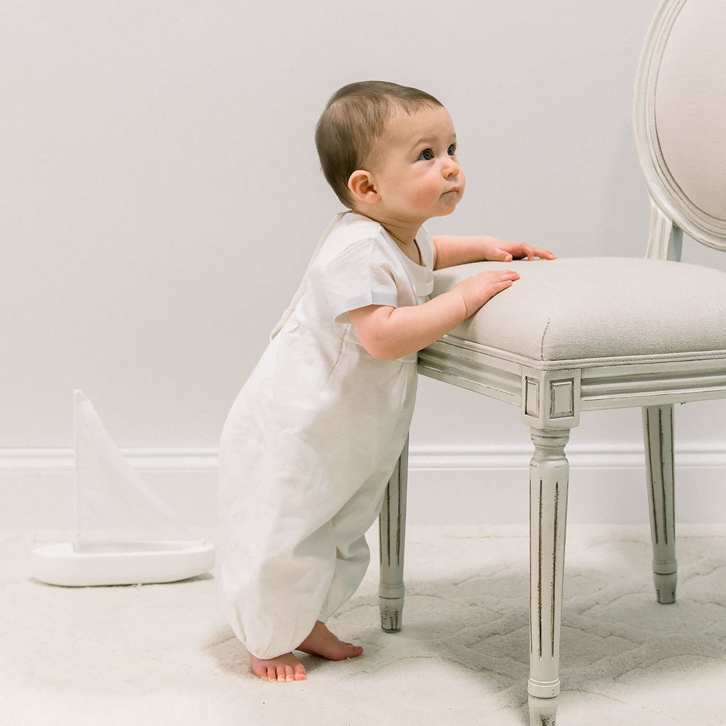 Baby boy standing up and wearing the Owen Linen Romper featuring silk ribbon in ivory and light blue across the front bodice and sleeves.